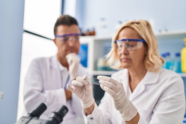 Middle age man and woman scientist partners holding sample working at laboratory - Foto, imagen
