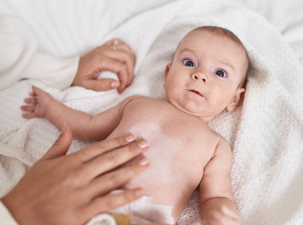Adorable caucasian baby lying on bed applying cream on body skin at bedroom - Foto, immagini