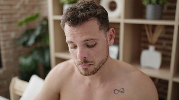 Young caucasian man thinking sitting on bed shirtless at bedroom - 写真・画像