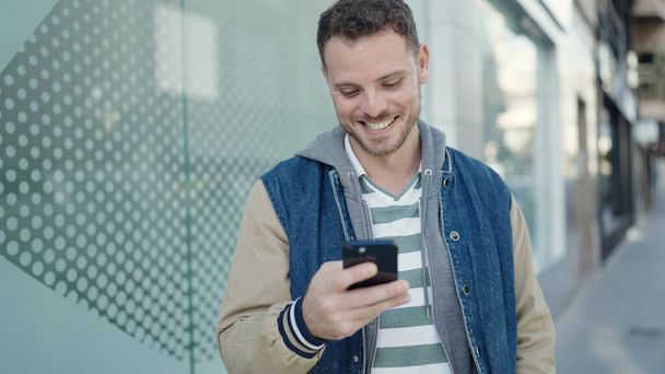 Young caucasian man smiling using smartphone at street - 写真・画像