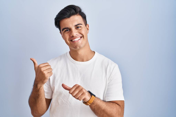 Homem hispânico em pé sobre fundo azul apontando para as costas com a mão e os polegares para cima, sorrindo confiante  - Foto, Imagem