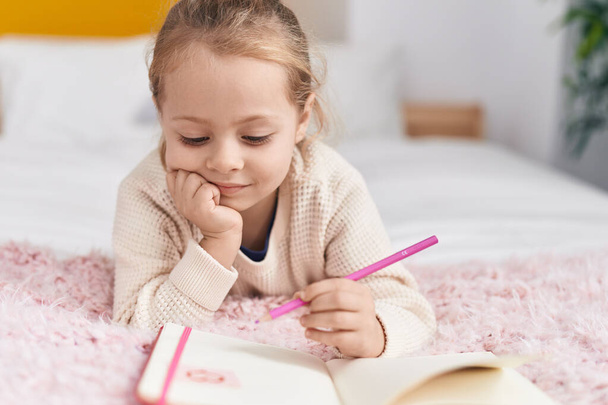 Schattig blond meisje tekening op notebook liggend op bed in de slaapkamer - Foto, afbeelding
