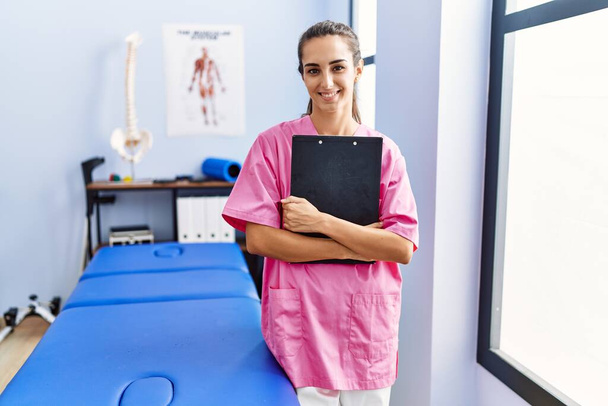 Young hispanic woman wearing physiotherapist uniform holding clipboard at physiotherpy clinic - 写真・画像