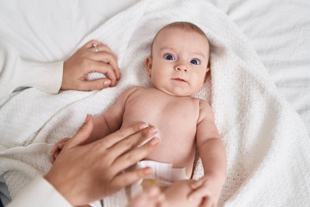 Adorable caucasian baby lying on bed applying cream on body skin at bedroom - Foto, afbeelding