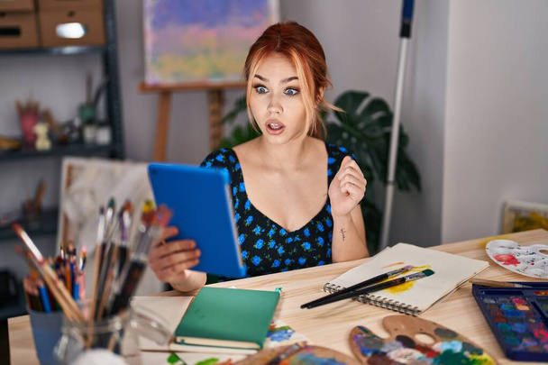 Young caucasian woman sitting at art studio doing video call with tablet scared and amazed with open mouth for surprise, disbelief face  - Foto, imagen