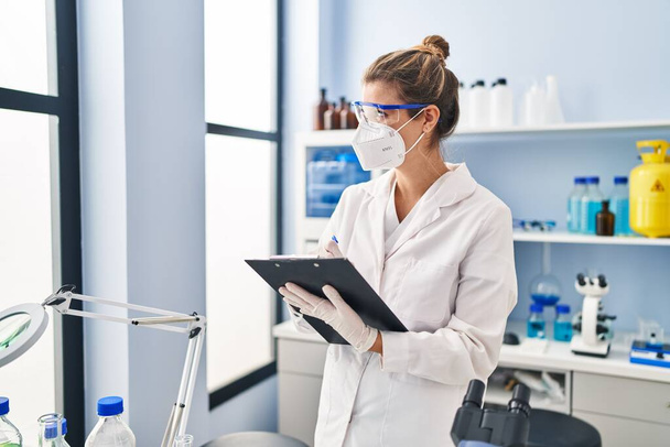 Junge blonde Frau in Wissenschaftleruniform und medizinischer Maske mit Schrift auf Klemmbrett im Labor - Foto, Bild