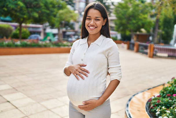 jong latin vrouw zwanger glimlachen zelfverzekerd aanraken buik in park - Foto, afbeelding