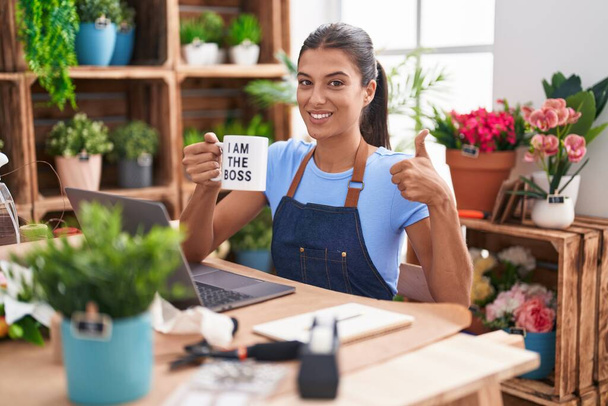 Brunette young woman working at florist shop holding i am the boss cup smiling happy and positive, thumb up doing excellent and approval sign  - Valokuva, kuva