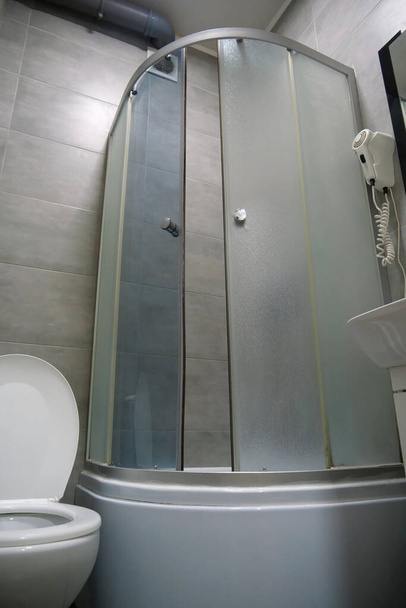 Intérieur de la salle de toilettes avec cuvette de toilette blanche avec couvercle ouvert, cabine de douche, lavabo, sèche-cheveux, armoire sous le lavabo et carreaux muraux et de sol gris - Photo, image