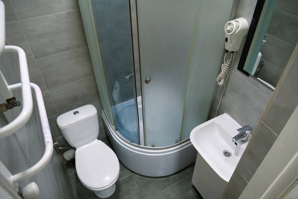 Toilettenraumausstattung mit weißer Toilettenschüssel, Duschkabine, Waschbecken, Fön, Handtuchheizung, Spiegel, Schrank unter dem Waschbecken und grauen Wand- und Bodenfliesen - Foto, Bild