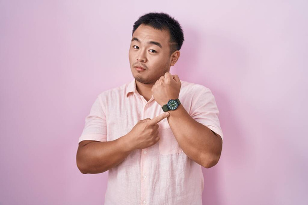 Chinese young man standing over pink background in hurry pointing to watch time, impatience, looking at the camera with relaxed expression  - Valokuva, kuva