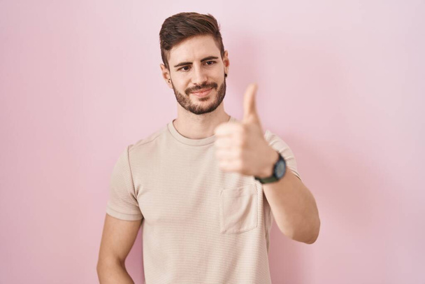 Hispanic man with beard standing over pink background looking proud, smiling doing thumbs up gesture to the side  - Foto, Imagen