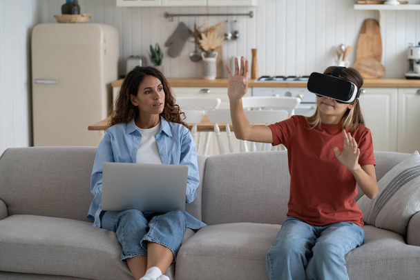 Interested teenage girl uses VR headset to visit metaverse or watch 3D virtual reality movies from comfort home. Progressive European schoolgirl waving hands sits on sofa near mother woman with laptop - Photo, Image