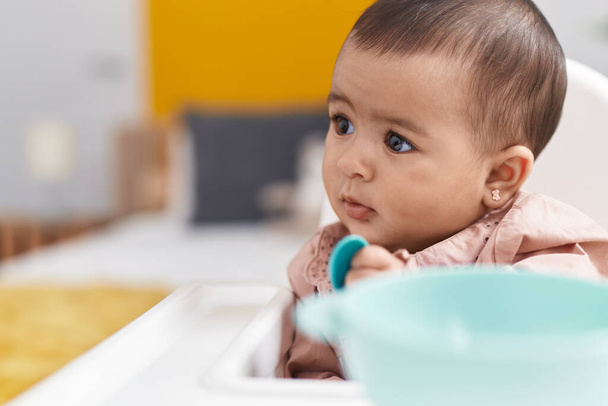 Adorable hispanic baby sitting on highchair holding spoon at bedroom - Photo, image