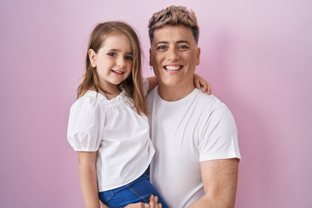 Νεαρός πατέρας αγκαλιάζει την κόρη πάνω από ροζ φόντο φαίνεται θετική και ευτυχισμένη στέκεται και χαμογελά με αυτοπεποίθηση χαμόγελο δείχνει τα δόντια  - Φωτογραφία, εικόνα