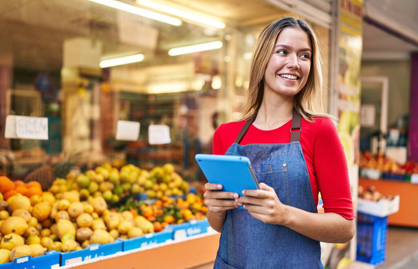 Young hispanic woman shop assistant using touchpad at fruit market - 写真・画像