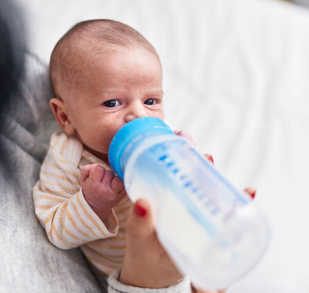 Adorable caucasian baby on mother arms sucking feeding bottle at bedroom - Photo, Image