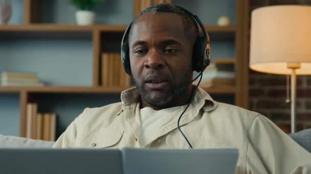 Mature african american man in headphones remote discuss with partner at online conference use laptop at home businessman talk on video call holding documents virtual negotiation concept distance work - Materiał filmowy, wideo