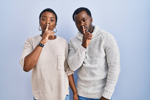 Ein junges afrikanisch-amerikanisches Paar steht vor blauem Hintergrund und bittet um Ruhe mit dem Finger auf den Lippen. Schweigen und geheimes Konzept.  - Foto, Bild