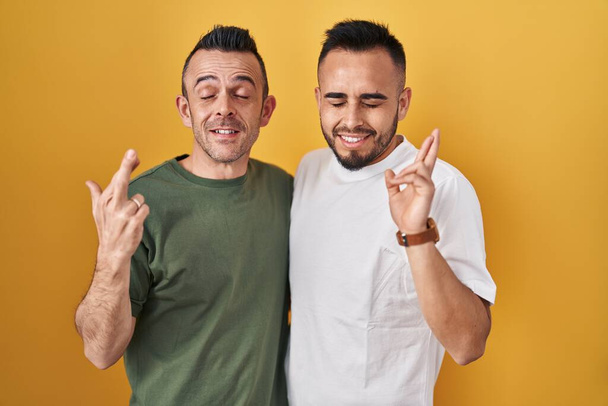 Homosexual couple standing over yellow background gesturing finger crossed smiling with hope and eyes closed. luck and superstitious concept.  - Foto, Bild