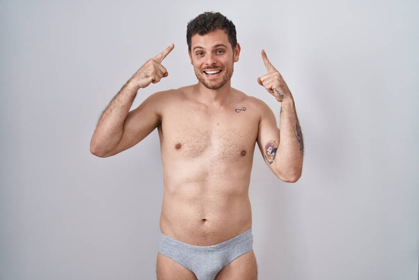 Νεαρός Ισπανός που στέκεται γυμνός φορώντας underware χαμογελώντας δείχνοντας το κεφάλι με τα δύο χέρια δάχτυλο, μεγάλη ιδέα ή σκέψη, καλή μνήμη  - Φωτογραφία, εικόνα