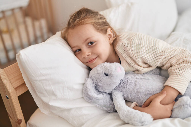Чарівна блондинка обіймає ляльку кролика, лежачи на ліжку в спальні
 - Фото, зображення