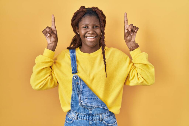 指で指差して腕を上げたアフリカの女性は、黄色の背景の上に立って笑顔と驚きと指摘.  - 写真・画像