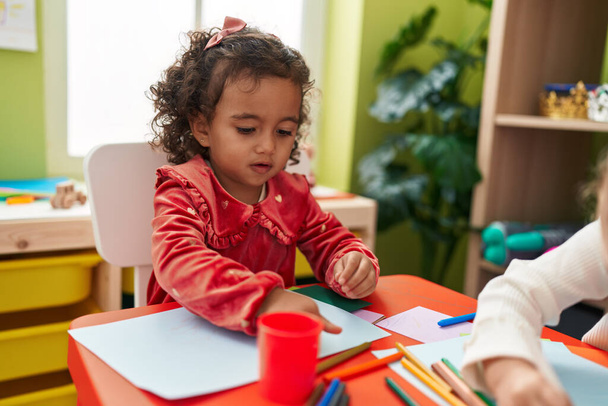 Чарівна іспаномовна дівчина дошкільного віку сидить на столі малює на папері в дитячому садку
 - Фото, зображення