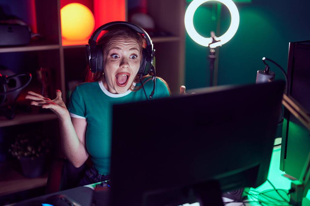 Redhead woman playing video games celebrating victory with happy smile and winner expression with raised hands  - Foto, Imagem
