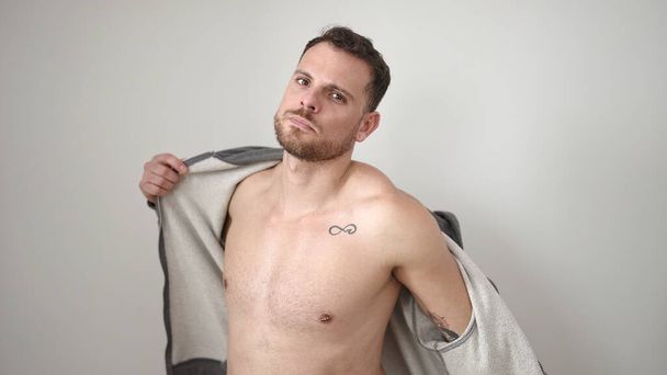 Young caucasian man standing with serious expression taking sweatshirt off over isolated white background - Fotó, kép