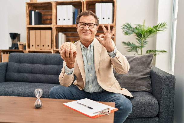 Homem psiquiatra sênior trabalhando no consultório fazendo ok assinar com os dedos, sorrindo amigável gesto excelente símbolo  - Foto, Imagem