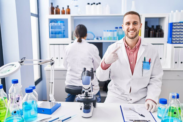 Fiatal férfi dolgozik tudós laboratóriumban mosolyogva boldog mutogatás kézzel és ujjal  - Fotó, kép