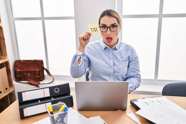 Młoda latynoska kobieta biznesu w biurze obliczając podatki przestraszony i zdumiony z otwartymi ustami dla zaskoczenia, niedowierzanie twarz  - Zdjęcie, obraz