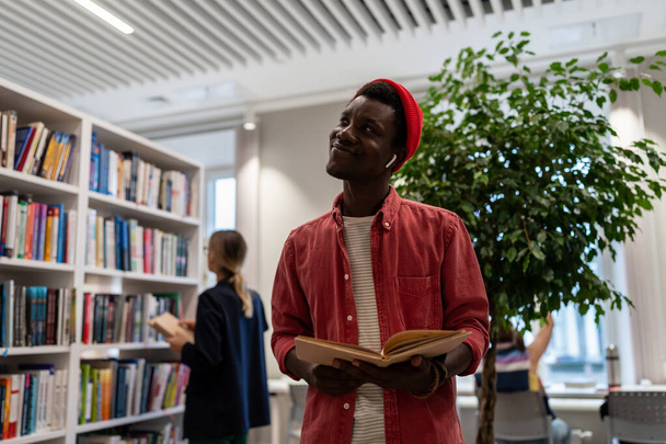 Ικανοποιημένος Αφροαμερικανός φοιτητής στέκεται στη βιβλιοθήκη με ανοιχτό βιβλίο φαίνεται όνειρο για πέρασε εξετάσεις απολαύσετε ενδιαφέρουσες αποτελεσματικές σπουδές στο ανώτερο σχολείο. Επιτυχημένος άριστος μαθητής παίρνει υποτροφία - Φωτογραφία, εικόνα