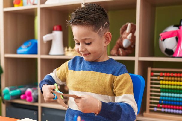 Adorable hispanic boy student smiling confident cutting paper at kindergarten - 写真・画像
