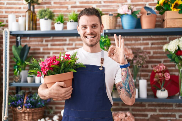 Young hispanic man working at florist shop holding plant pot doing ok sign with fingers, smiling friendly gesturing excellent symbol  - Фото, изображение