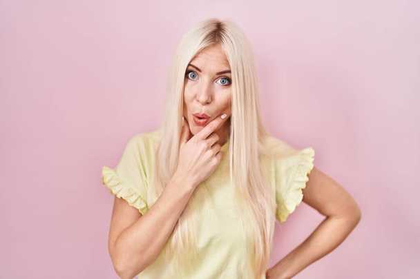 Caucasian woman standing over pink background looking fascinated with disbelief, surprise and amazed expression with hands on chin  - Foto, afbeelding