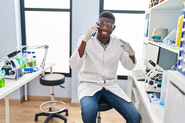 Uomo afroamericano che lavora presso il laboratorio scientifico parlando al telefono sorridendo felice indicando con mano e dito  - Foto, immagini