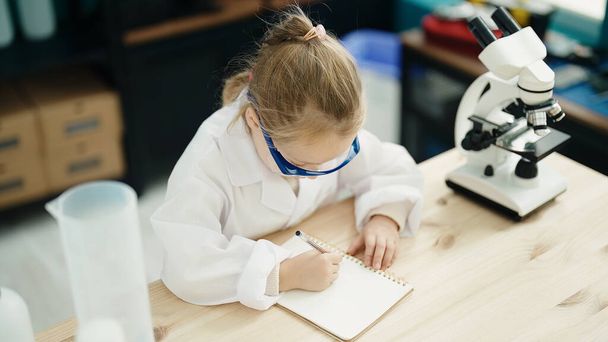 Adorable blonde girl student using microscope writing on notebook at laboratory classroom - Photo, Image