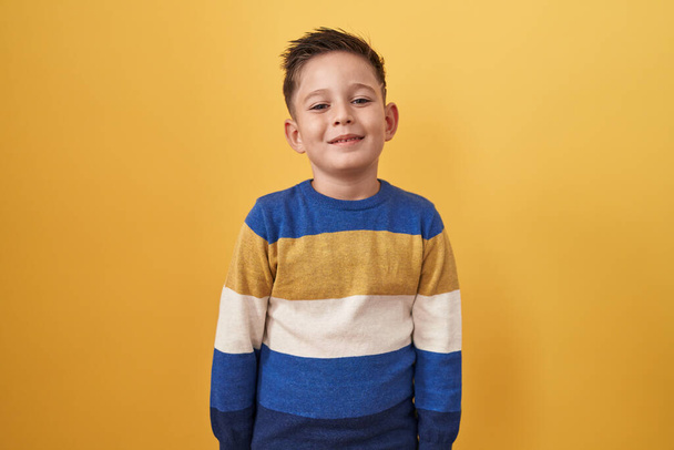 Mały latynoski chłopiec stojący na żółtym tle ze szczęśliwym i chłodnym uśmiechem na twarzy. szczęśliwy człowiek.  - Zdjęcie, obraz