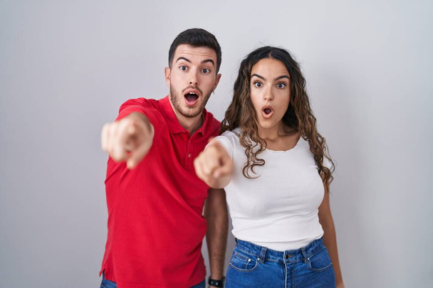 Young hispanic couple standing over isolated background pointing with finger surprised ahead, open mouth amazed expression, something on the front  - Foto, immagini