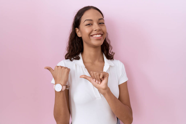 Jonge Spaanse vrouw draagt casual witte t-shirt wijzen naar de achterkant met de hand en duimen omhoog, glimlachen vol vertrouwen  - Foto, afbeelding