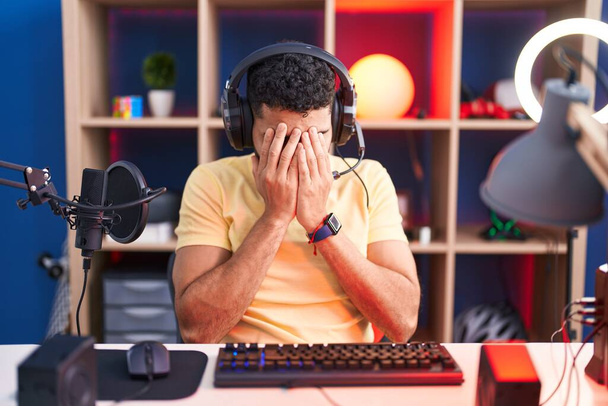Латиноамериканец с бородой играет в видеоигры с наушниками с грустным лицом, прикрывающим лицо руками во время плача. депрессия.  - Фото, изображение