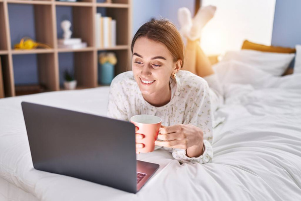 Junge kaukasische Frau benutzt Laptop und trinkt Kaffee im Schlafzimmer - Foto, Bild