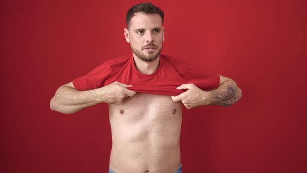 Young caucasian man standing with serious expression taking t shirt off over isolated red background - Fotó, kép