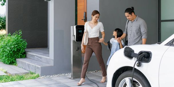 Progressive young parents and daughter with electric vehicle and home charging station. Green and clean energy from electric vehicles for healthy environment. Eco power from renewable source at home. - 写真・画像