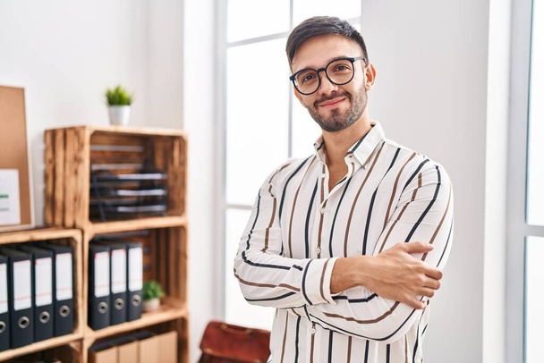 Молодой латиноамериканец-бизнесмен улыбается уверенно стоя со скрещенными руками в офисе - Фото, изображение