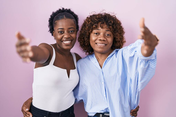 Two african women standing over pink background looking at the camera smiling with open arms for hug. cheerful expression embracing happiness.  - Photo, Image