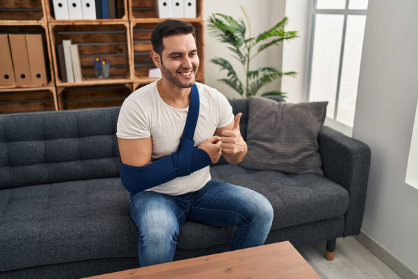 Young hispanic man with beard wearing arm on sling sitting at therapy consult smiling happy and positive, thumb up doing excellent and approval sign  - Photo, image