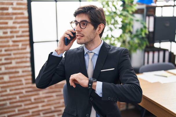 Young hispanic man business worker talking on smartphone looking watch at office - Photo, Image